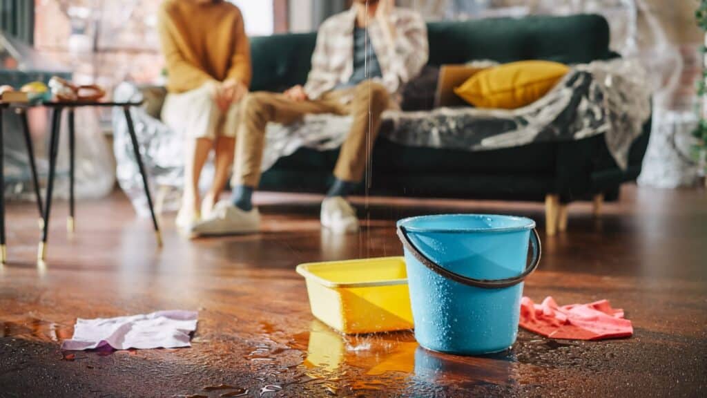 Two people sit on a couch wrapped in plastic with buckets on the ground collecting water from a leaking ceiling.