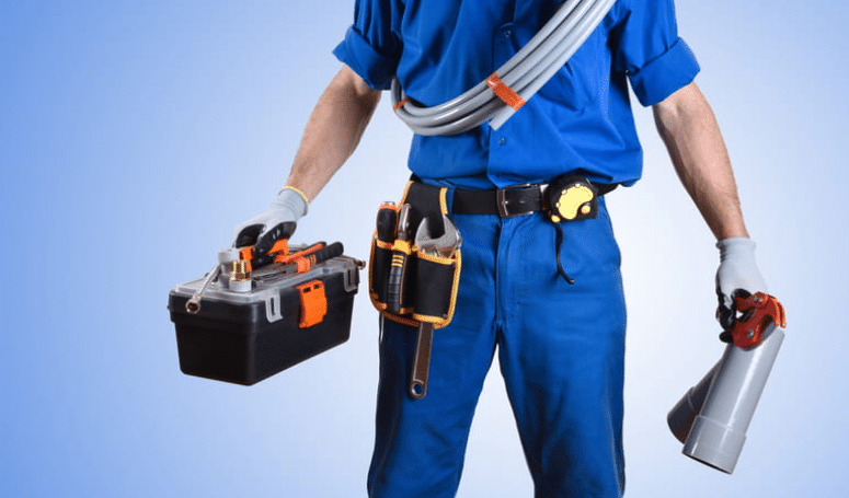 A plumber holding tools and pipes with additional plumbing tools strapped to him. 