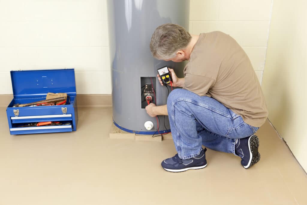 plumber kneeling down and repairing water heater using common plumbing tools including an analog multimeter.